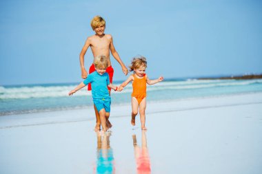 Group of three children boys and girl run on sand beach holding hands wear swimsuits during hot summer day