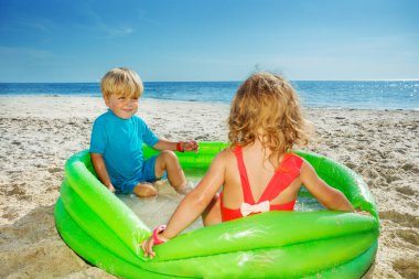 Two happy kids play splashing in the inflatable pool at the sea beach looking at each other over waves on background