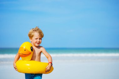 Happy little blond boy with inflatable yellow duck buoy stand on the sand beach close-up smiling portrait