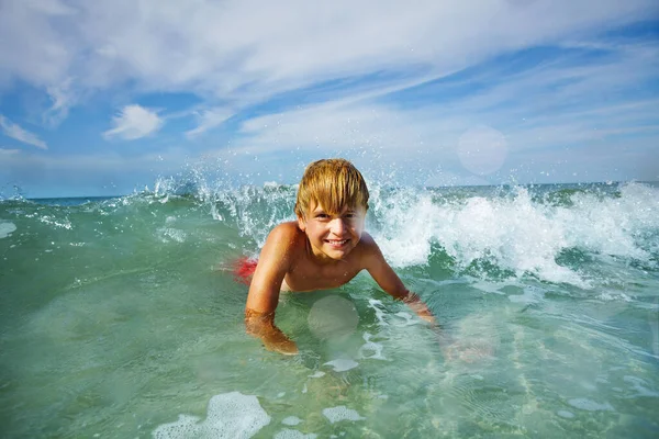 Happy smiling young blond boy lay in ocean waves on the beach with water splashes and drops flying around