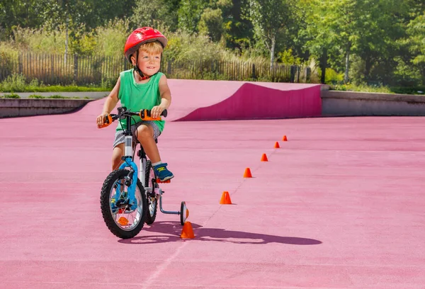Little blond boy ride small bicycle with learning wheels roll around orange cones on color surface of the park