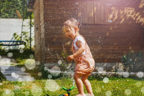 Happy beautiful girl in wet clothes play with water sprinkler in the garden lawn on hot summer day