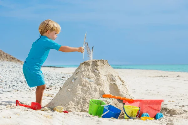 Handsome little boy building sand castle putting feather on top of the tower play on white ocean beach