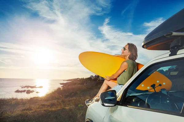 Young woman with surfboard sit on a car hood look over sea and sunset