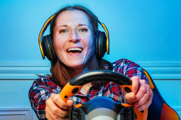Happy gamer woman in headphones with hands on console steering wheel of a laughing playing race game