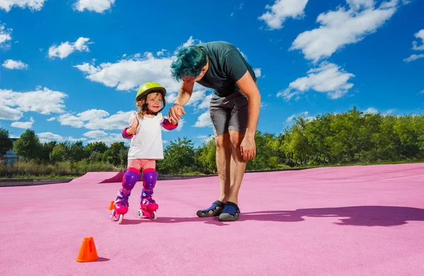 Father teach little happy girl to stand on rollers holding her with hands at the color skate park