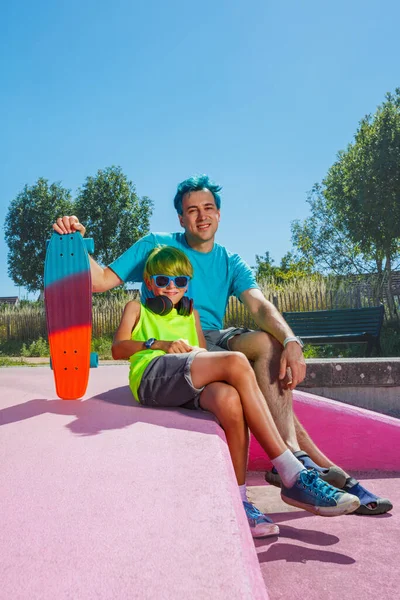 Blue hair adult man sit with a boy at skatepark holding skateboard laughing together