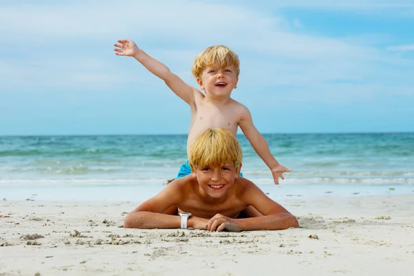 Two smiling boys with younger brother sit on on top of another and lift up hands at the sea sand beach at summer vacation