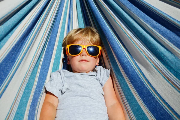 Close-up view from above of a cute calm little blond boy in sunglasses rest in hammock