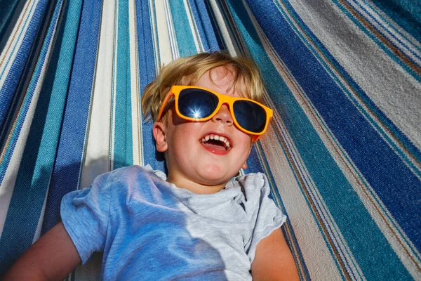 Close-up view from above of a laughing cute little blond boy in sunglasses rest in hammock