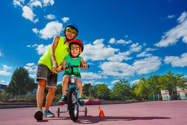 Preteen boy help little brother to learn ride a bicycle holding the bike with hands in the park
