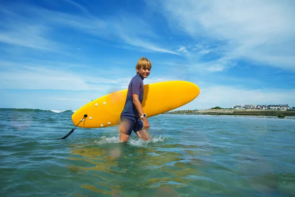 Cute young boy in swimsuit walk holding orange surfboard in waves and smile