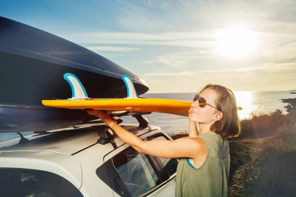 Young surfer woman take surfboard from roof top rack baggage on the car over ocean sunset view