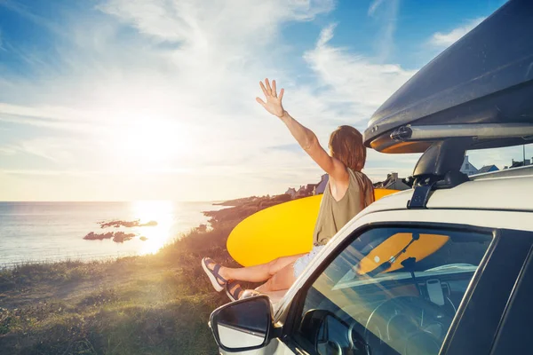 Young woman with surfboard sit on a car hood look over sea and sunset lift one hand happily up