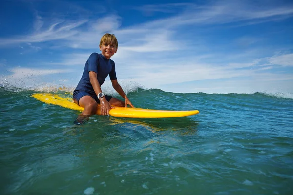 Cute smiling boy sit on the surf board looking at camera in the sea in waves with splashes