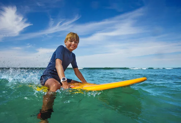 Portrait of smiling boy sit on the surf board looking at camera in the sea learning at surfing school