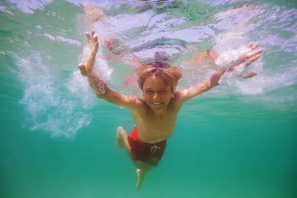 Happy smiling boy swim underwater in the ocean with open eyes and posing over sunlight lit background
