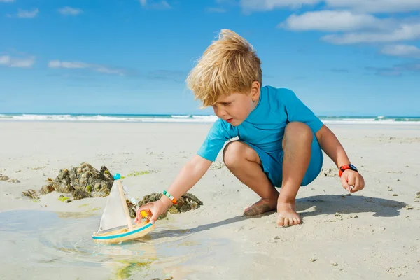Cute handsome little blond boy put toy boat in the paddle waves at the sea beach during summer vacation