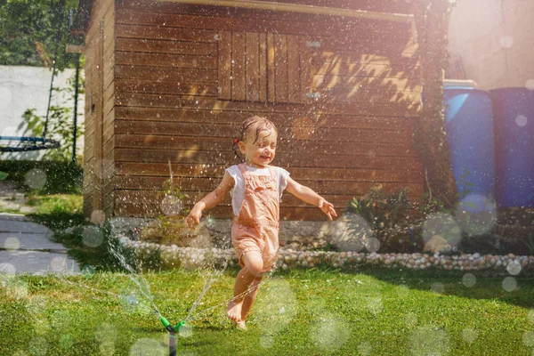 Happy beautiful girl in wet clothes run around with water sprinkler in the garden lawn on hot summer day