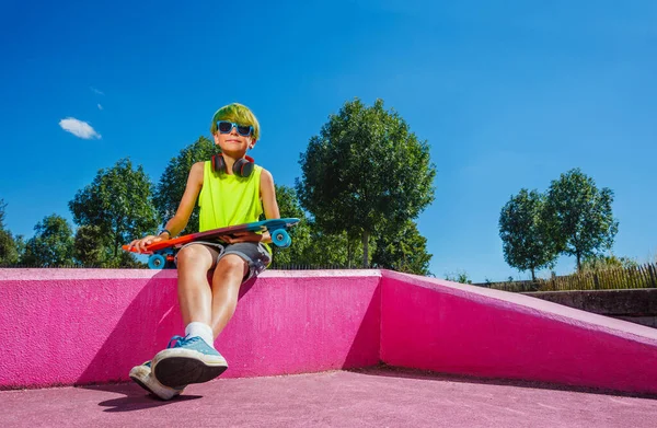 Low angle portrait of a cute young preteen boy with green hair sit on ramp holding skateboard in city skatepark