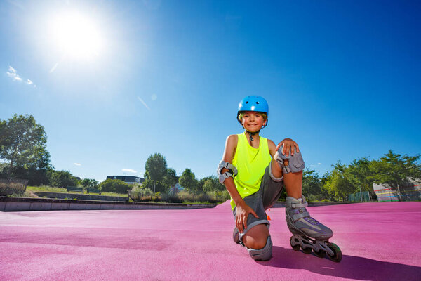 Happy preteen boy sit and pose in blue helmet with rollerblades in the skatepark wearing protection