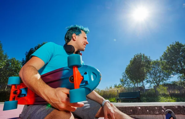 Man with blue hair sit on the pink ramp at the skatepark holding skate board over sun in the sky