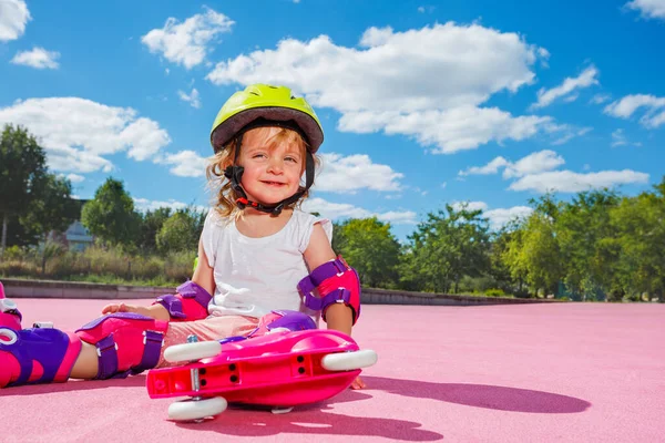 Smiling little girl learn to skate on rollers, sit with helmet and protection on the color floor of the skatepark