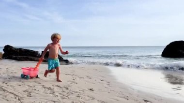 Happy little Caucasian blond boy run with toy  on beach over ocean waves