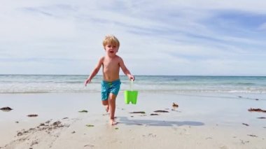 Happy little Caucasian blond boy run with plastic toy bucket on the white sand beach over ocean waves