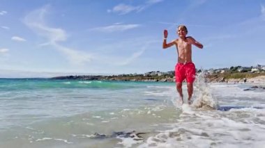 Happy little boy stand and jump in ocean waves at the sand beach on the summer vacation