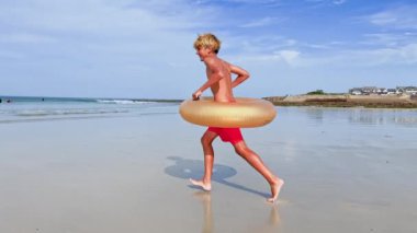 Şişme donut şamandıralı mutlu yakışıklı çocuk kumsalda gülümsüyor.