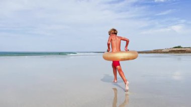 Happy handsome boy with inflatable doughnut buoy run smiling on the sand beach