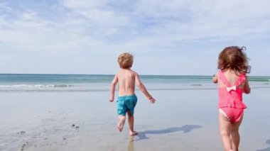 Two happy little kids  run on the sand beach in ocean waves on summer vacation 