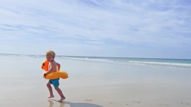 Happy little blond boy with inflatable yellow duck buoy run on the sand beach