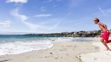 Happy little boy run and jump into ocean wave on the sand beach on the summer vacation