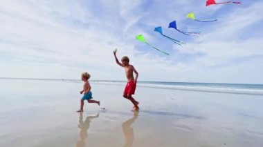 Two handsome brother boys run together holding colorful  kite over blue sky at the ocean beach having fun on summer vacation