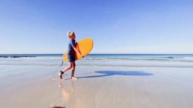 Cute young boy in swimsuit walk holding orange surfboard at sand ocean beach