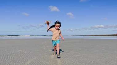 Kafkasyalı şirin sarışın çocuk pilot kafasında uçak oyuncağıyla koşuyor ve beyaz kumsalda deniz dalgaları üzerinde google arıyor.