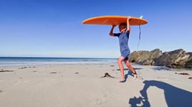 Cute young boy in swimsuit walk holding orange surfboard at sand ocean beach