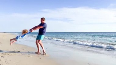 Baba oğluyla kumsalda oynar el ele döner ve etrafında döner.