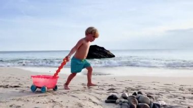 Happy little Caucasian blond boy run with toy  on beach over ocean waves
