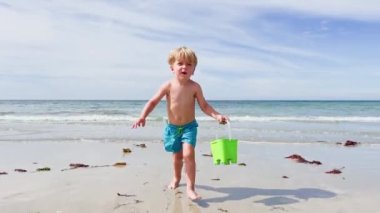 Happy little Caucasian blond boy run with plastic toy bucket on the white sand beach over ocean waves