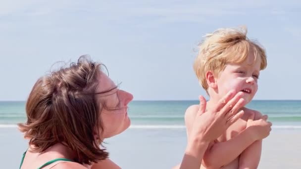 海滩上快乐的妈妈在暑假给可爱的金发碧眼的小男孩涂防晒霜 — 图库视频影像