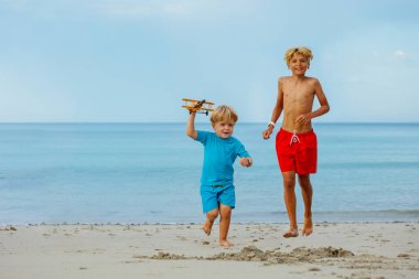 Little boy with older brother run holding toy model of the plane over calm ocean and beach on background