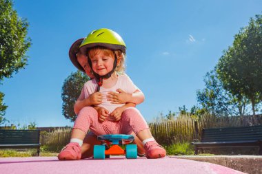 Two little children girl with brother boy sit on the skate playing at skatepark wearing helmets on sunny summer day