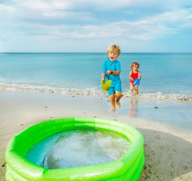 Two happy kids run to inflatable pool bringing water from sea in buckets and splash inside