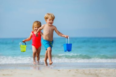 Little boy and a girl, siblings run barefoot on sea beach from waves carry toy buckets with water