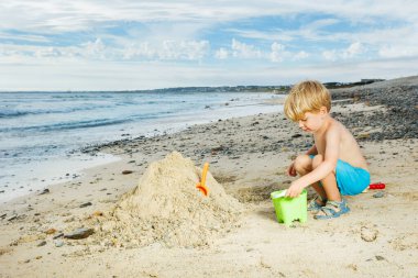 Happy little blond boy play with sand building sandcastle on the sea beach during vacation