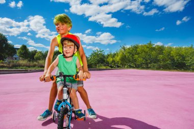 Two brothers together portrait in action, big boy push little bicycle teaching smaller kid to ride
