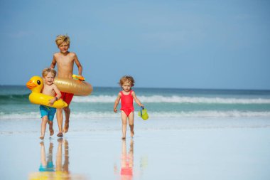 Three cute kids boys with inflatable toy buoys doughnut and duck having fun running on the sand beach